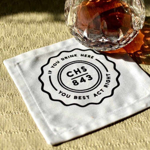 Charleston cocktail napkin with "If you drink here you best act right" design next to a glass on textured surface